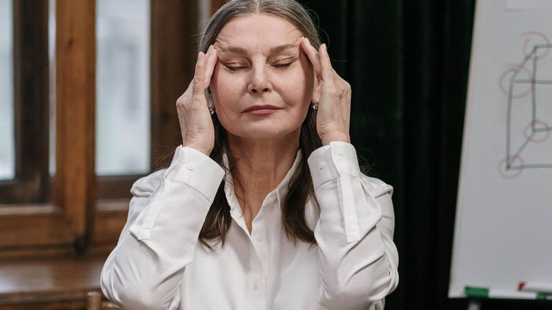 Woman in her fifties pressing her fingertips to her temples, eyes closed, struggling with brain fog